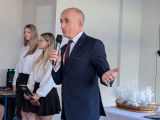 Mężczyzna w ciemnym garniturze mówi przez mikrofon gestykulując. Obok dwie długowłose uczennice w białych bluzkach