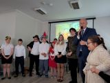 Mężczyzna w ciemnym garniturze mówi przez mikrofon. Obok grupa uczniów w przebraniach postaci z bajek i strojach galowych