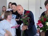Mężczyzna w ciemnym garniturze, z różą w dłoni pochyla się nad uśmiechniętą uczennicą w białej bluzce