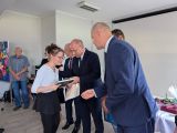Trzech mężczyzn w granatowych garniturach. Obok dziewczyna w okularach i białej bluzce ze świadectwem w dłoni