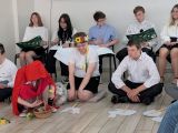 Uśmiechnięta trójka uczniów siedząca po turecku. Za nimi grupa uczniów siedząca na krzesłach