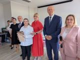 Uśmiechnięta uczennica w białej bluzce ze świadectwem w dłoni. Obok niej grupa uśmiechniętych nauczycieli