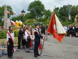 Na pierwszym planie troje nastolatków ze sztandarem szkoły. W tle Grupa kilkunastu innych osób i pomnik Łokietka.