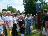Przed małą, przydrożną kapliczką stoi grupa osób i słuch stojącego przed nimi mężczyznę.
