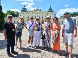 Na placu przed kremowym pałacem stoi pieć kobiet w przeciwsłonecznych okulaarach i dwóch mężczyzn.