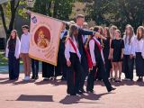 Troje uczniów w strojach galowych maszeruje na dziedzińcu szkoły ze sztandarem. W tle grupa uczniów w strojach galowych