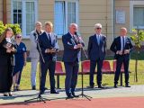Mężczyzna w granatowym garniturze przemawia przez mikrofon na dziedzińcu szkoły. W tle grupa ludzi w strojach galowych