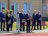 Szczupły mężczyzna w ciemnym garniturze, z teczką w dłoni przemawia przez mikrofon. W tle grupa ludzi w strojach galowych