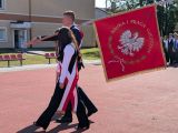 Uczeń w garniturze maszeruje po dziedzińcu szkoły ze sztandarem na ramieniu. Obok niego dwie uczennice w strojach galowych