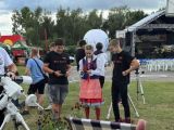 Dziewczyna ubrana w ludowy strój trzyma sterownik do robotów, po bokach stoi dwóch uczniów. Jeden trzyma sterownik, drugi...