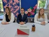 Uczeń w garniturze i dwie dziewczyny w białych bluzkach siedzą za białym stołem. Przed nimi biało-czerwona urna wyborcza