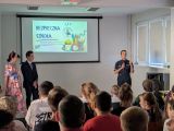 Uczniowie siedzący na krzesłach w dużej sali. Przed nimi policjantka, kobieta w kolorowej sukni j chłopak w garniturze. W...