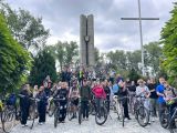 Duża grupa młodzieży stoi przy rowerach. Za nimi betonowy pomnik.