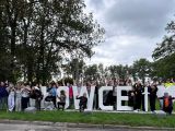 Duża grupa młodzieży szkolnej stoi między betonowymi, dużymi literami, które tworzą napis Płowce". Wokół drzewa.