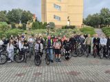 Duża grupa uczniów liceum stoi na dziedzińcu szkoły przy rowerach. W tle pomarańczowy budynek.