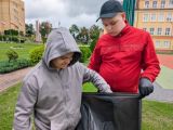 Uśmiechnięty uczeń w szarej bluzie wrzuca odpad do szarego foliowego worka trzymanego przez chłopca w czerwonej bluzie