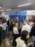 Zdjęcie przedstawia grupę ludzi rozmawiających i przeglądających materiały przy stoiskach promocyjnych. Stoiska są...
