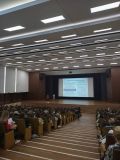 Zdjęcie przedstawia dużą salę wykładową. Wiele rzędów drewnianych krzeseł jest wypełnionych ludźmi, którzy słuchają...