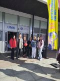 Grupa siedmiu osób stoi na zewnątrz budynku z tablicą UMK (Uniwersytet Mikołaja Kopernika) nad wejściem. Wszyscy uśmiechają...