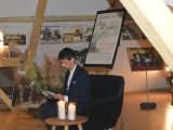 Uczeń liceum w garniturze siedzi w fotelu i czyta książkę. Przed nim na stoliku palą się dwie świece. W tle okazały bukiet...
