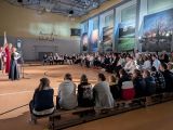 Grupa uczniów siedząca na ławeczkach w sali gimnastycznej. Po lewej uczennice przemawiające przez mikrofon