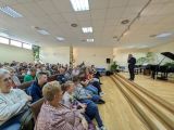 Duża grupa ludzi siedzi na krzesłach w dużej sali. Przed nimi na scenie mężczyzna w garniturze z mikrofonem w dłoni