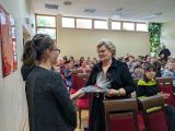 Dwie dziewczyny wręczają upominek zapakowany w folię starszej kobiecie ubranej na czarno. W tle siedząca na krzesłach duża...