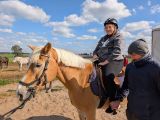 Uczennica w okularach i kasku na głowie siedzi na jasnym koniu. Obok mężczyzna w czapce