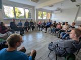 Duża grupa uczniów siedzi na krzesłach tworząc okrąg w dużej, jasnej sali. Między nimi mężczyzna w niebieskim swetrze