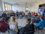 Duża grupa uczniów siedzi na szarych krzesłach w dużej, jasnej sali. Między nimi kobieta w czerwonej bluzie