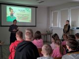 Kobieta w czarnej koszuli mówi do młodzieży siedzącej w dużej sali. Obok młoda kobieta w futrzanym bezrękawniku. Za nimi...