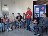 Kobieta w czerwonej bluzie i jeansach żywo gestykuluje stojąc. Obok niej grupa młodzieży siedząca na krzesłach