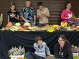 Długi stół przykryty czarnym obrusem. Na stole kolorowe dynie, kalafiory śliwki i inne jesienne owoce i warzywa. Przed...