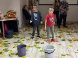Dwóch chłopców wrzuca z odległości ziemniaki do siwych wiaderek. Na podłodze porozrzucane suche liście. Po lewej na stole...