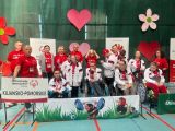 Grupa ludzi w sportowych biało-czerwonych dresach. Z przodu dzieci i młodzież na wózkach inwalidzkich. W tle powieszone...