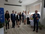 Mężczyzna w garniturze z jabłkiem w dłoni mówi przez mikrofon. Za nim grupa uczniów ubranych w galowe stroje