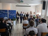 Duża grupa uczniów siedząca w na krzesłach w jasnym pomieszczeniu. Z przodu uczniowie w strojach galowych ustawieni w rzędzie