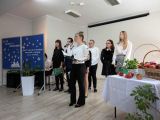 Dziewczyna z kokiem na głowie, w stroju galowym śpiewa przez mikrofon. Za nią grupa uczniów w strojach galowych