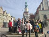 Grupa uczniów w jesiennych kurtkach stoi przed fontanną. W tle wieża ratusza miejskiego. Po prawej i lewej kamienice