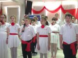 Grupa dzieci w strojach galowych z czerwonymi szarfami i kotylionami śpiewa stojąc w trzech rzędach