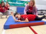 Chłopiec w okularach turla się po kolorowym materacu z pomocą uśmiechniętej nauczycielki