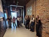 Kilkanaście uczennic liceum stoi na korytarzu muzealnym. Z prawej strony ściana z cegieł. Wszystkie mają słuchawki na uszach.