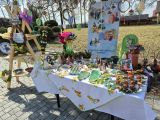 Długi stół z dużą ilością ceramicznych i krawieckich wytworów o tematyce wielkanocnej. Za stołem banner reklamowy szkoły