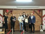 Na scenie stoi siedem osób podczas uroczystego wręczenia nagród. W centrum znajduje się młoda laureatka trzymająca dyplom i...