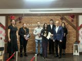 Na scenie stoi siedem osób w eleganckiej, uroczystej atmosferze. w centrum młoda dziewczyna prezentuje statuetkę i dyplom,...