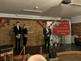 Na scenie stoją trzy elegancko ubrane osoby na tle ceglanej ściany ozdobionej czerwonymi sercami, obok dużego serca z...