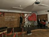 Na scenie z ceglaną ścianą i drewnianą podłogą. Po lewej stronie uczestniczka śpiewa do mikrofonu, a po prawej widnieje...