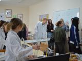 W sali chemicznej trzy osoby w białych fartuchach wykonują doświadczenia chemiczne. Kilka osób się temu przygląda.