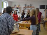 Kilkanaście osób stoi wokół ławek szkolnych. Wszyscy trzymają się za ręce. Na stolikach przyrządy fizyczne.