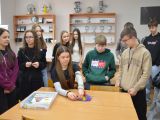 Przt stoliku siedzi licealistka i pokazuje urządzenie fizyczne. Za nią stoi kilkunastu uczniów szkoły podstawowej.